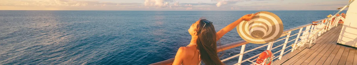 Frau mit Sonnenhut an der Reling eines Kreuzfahrtschiffes blickt bei Sonnenuntergang auf das offene Meer.