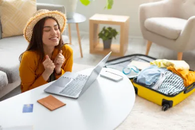 Junge Frau freut sich vor einem Laptop über ihre gerade gebuchte Urlaubsreise, neben ihr ein gepackter Koffer und ein Reisepass.