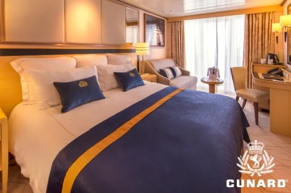 Britannia Club Balkonkabine auf der Queen Mary 2 Doppelbett mit weißen Bezügen und blauer Tagesdecke in einer Britannia Club Balkonkabine auf der Queen Mary 2. Im Hintergrund ein 2-sitziges Sofa, ein Schreibtisch mit Stuhl und die Tür zum Balkon mit Vorhängen.