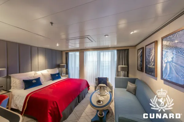 Princess Suite auf der Queen Mary 2 Großes Doppelbett mit weißen Bezügen, roter Tagesdecke & blauen Kissen in einer Princess Suite auf der Queen Mary 2. Rechts daneben großes Sofa mit Tisch und Sessel, im Hintergrund Balkontür mit zugezogenen Vorhängen.