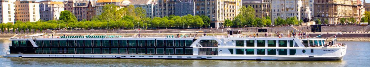 Das Flusskreuzfahrtschiff MS VistaNova von 1A Vista Reisen in voller Länge auf der Donau, mit dem Passagier-Sonnendeck und der Uferpromenade von Budapest im Hintergrund.