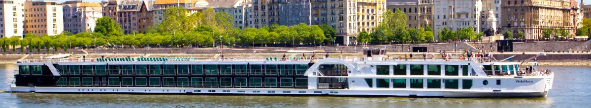 Das Flusskreuzfahrtschiff MS VistaNova von 1A Vista Reisen in voller Länge auf der Donau, mit dem Passagier-Sonnendeck und der Uferpromenade von Budapest im Hintergrund.