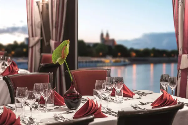 Elegant gedeckter Tisch im Bordrestaurant der MS VistaNova mit weißen Tischdecken und roten Servietten. Durch das Panoramafenster blickt man auf den Fluss und eine Stadt am Ufer in der Abenddämmerung.