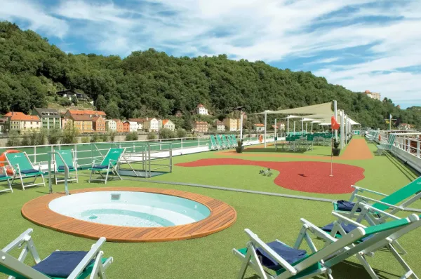 Das Sonnendeck der MS VistaNova mit einem runden Pool, Liegestühlen und einem Putting Green. Im Hintergrund ist eine Stadt am Flussufer und ein bewaldeter Hügel zu sehen.