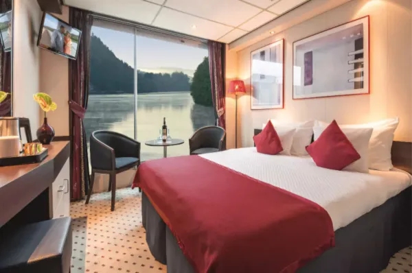 Modern eingerichtete Kabine auf der MS VistaNova mit Doppelbett, Schreibtisch und einem französischen Balkon, der den Blick auf eine abendliche Flusslandschaft freigibt.