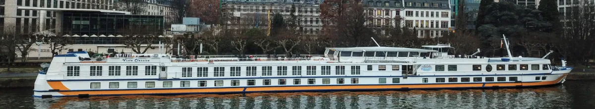 Das Flusskreuzfahrtschiff MS VistaClassica liegt am Abend an einem Kai vor einer modernen Stadtsilhouette, wobei sich die Lichter im Wasser spiegeln. Auf dem Bug des Schiffes ist ein kleiner, geschmückter Weihnachtsbaum zu erkennen.