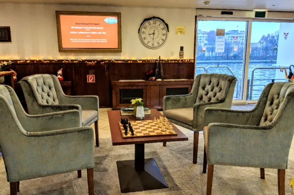 Lounge der MS VistaClassica mit Schachtisch Gemütliche Sitzecke in der Lounge der MS VistaClassica mit vier Sesseln um einen Schachtisch. Im Hintergrund ist die holzvertäfelte Wand und der Blick durch die Glastür nach draußen auf den Fluss zu sehen.