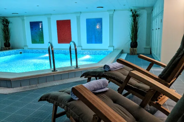Indoor-Pool der MS VistaClassica mit Ruheliegen Blick über zwei hölzerne Ruheliegen auf den beleuchteten Indoor-Pool der MS VistaClassica. An der Wand im Hintergrund befinden sich farbige Kunstpaneele und dekorative Pflanzen.