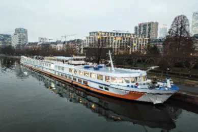 Das Flusskreuzfahrtschiff MS VistaClassica liegt bei Dämmerung an einer Uferpromenade. Die Lichter des Schiffes und der modernen Skyline im Hintergrund sind erleuchtet.
