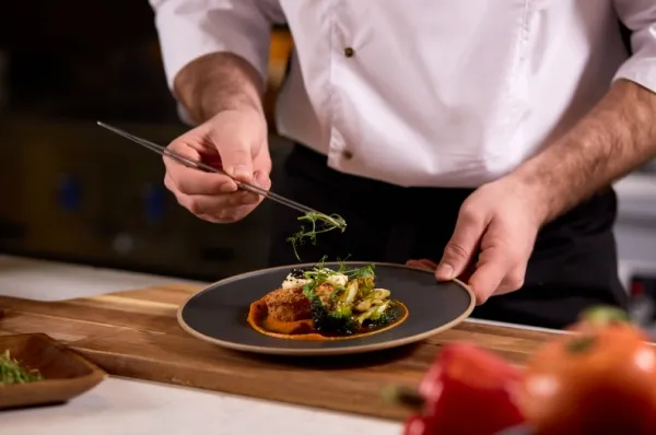 Ein Chefkoch richtet ein Gourmetmenü auf einem dunklen Teller an. Er trägt eine Schürze und frische Paprika liegen auf der Fläche.