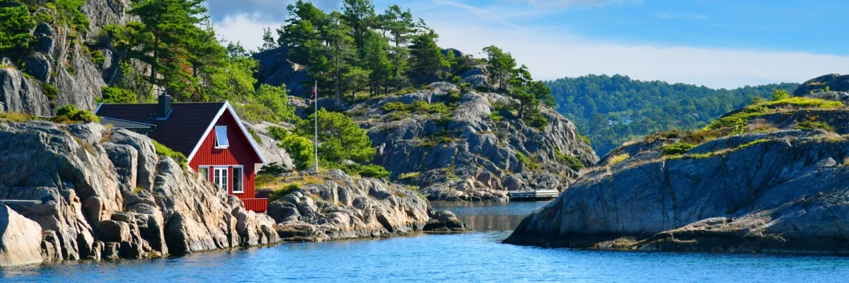 AIDA Herbst Kreuzfahrten nach Nordeuropa Idyllische Fjordlandschaft bei Kristiansand, Norwegen. Links ein rotes Holzhaus auf einem leicht bewaldeten Felsen, mittig und rechts weitere Felseninseln mit Gras und Bäumen.