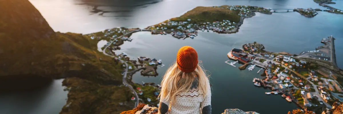 AIDA Nordland Kreuzfahrt – Spektakulärer Ausblick auf skandinavische Fjorde und Küsten im Herbst Frau in Strickpullover und roter Mütze blickt vom Berg auf eine skandinavische Schärenlandschaft und Küstenstadt.