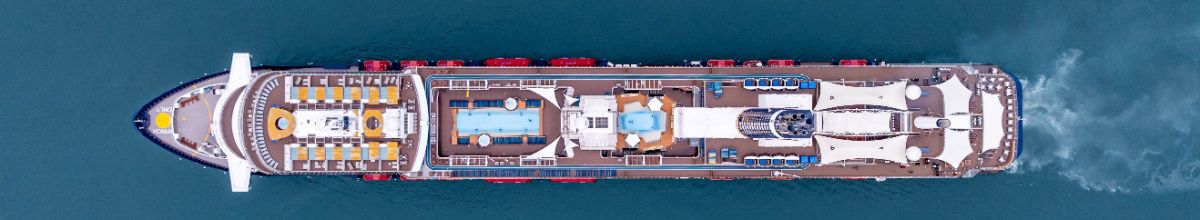 Vogelperspektive eines modernen Kreuzfahrtschiffes, das auf dem tiefblauen Meer fährt. Gut zu erkennen sind die Decks mit Pools, gelben und blauen Liegestühlen sowie das Kielwasser am Heck des Schiffes.