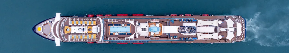 Luftaufnahme eines modernen Kreuzfahrtschiffes auf dem offenen Meer Vogelperspektive eines modernen Kreuzfahrtschiffes, das auf dem tiefblauen Meer fährt. Gut zu erkennen sind die Decks mit Pools, gelben und blauen Liegestühlen sowie das Kielwasser am Heck des Schiffes.