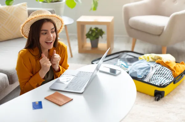 Junge Frau freut sich über eine gelungene Kreuzfahrt-Buchung am Laptop Junge Frau mit Strohhut sitzt begeistert vor ihrem Laptop auf dem Boden eines Wohnzimmers. Neben ihr liegt ein offener Koffer, ein Reisepass und eine Kreditkarte, was auf eine erfolgreiche Reisebuchung hindeutet.