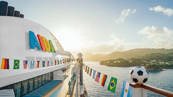 Deck eines AIDA-Kreuzfahrtschiffs mit internationalen Länderflaggen und einem Fußball im Vordergrund, im Hintergrund eine tropische Küstenlandschaft im Gegenlicht