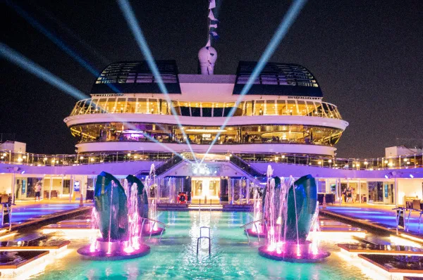 Das hell erleuchtete Hauptpooldeck der MSC Virtuosa bei Nacht, mit dem Atmosphere Pool und farbigen Wasserfontänen im Vordergrund und Scheinwerfern, die vom Schiff in den Himmel strahlen.