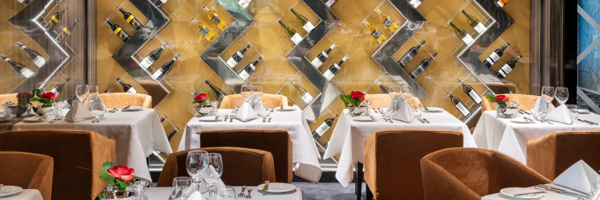 Ein elegantes Restaurant auf der MSC Virtuosa mit festlich gedeckten Tischen, bequemen braunen Stühlen und einer auffälligen Wanddekoration mit Weinregalen und Flaschen.