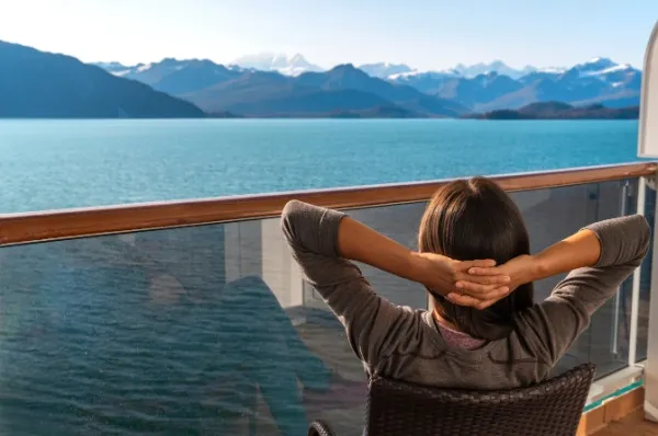 Frau sitzt auf Balkon eines Kreuzfahrtschiffes und hat die Arme hinter dem Kopf verschränkt, dabei guckt sie in die Ferne auf eine Berglandschaft.