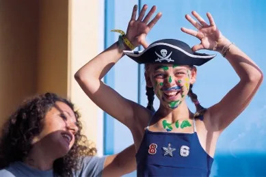 Ein Mädchen trägt einen Piratenhut und ist bunt angemalt und hebt die Hände in die Luft während sie in die Kamera lacht. Im Hintergrund ist eine Frau.