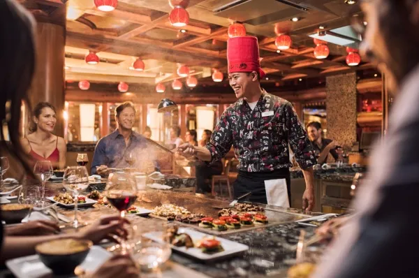 Ein Koch bereitet Essen in einer heißen Pfanne zu während Personen um ihn herum sitzen und zuschauen.