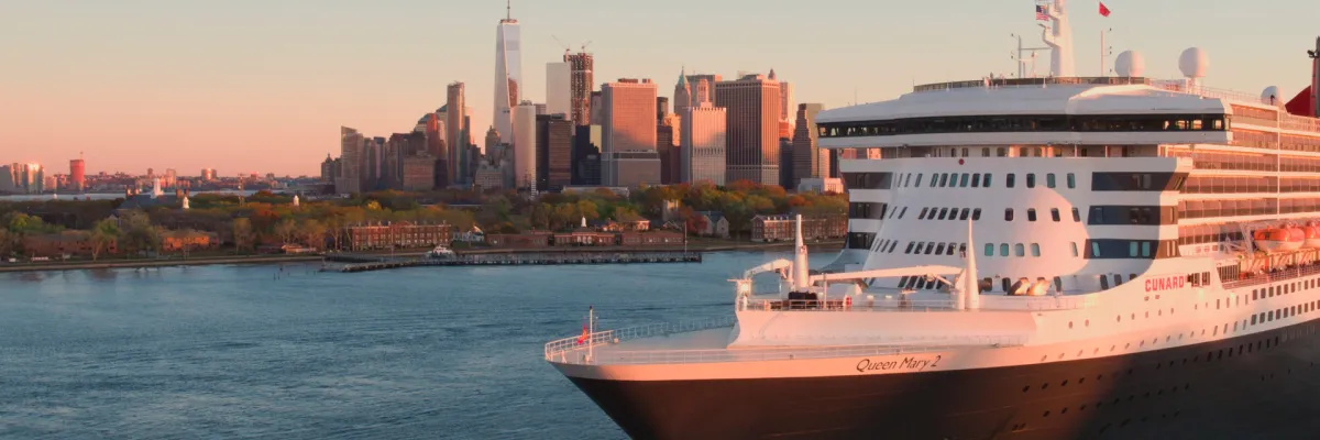 Das Kreuzfahrtschiff Queen Mary 2 von Cunard fährt bei Sonnenaufgang im Hafen von New York, mit der Skyline von Manhattan im Hintergrund.