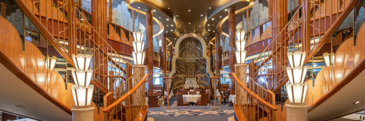 Das prunkvolle, mehrstöckige Britannia Restaurant auf einem Cunard-Schiff, dominiert von einer eleganten Holztreppe und leuchtenden Art-déco-Lampen.