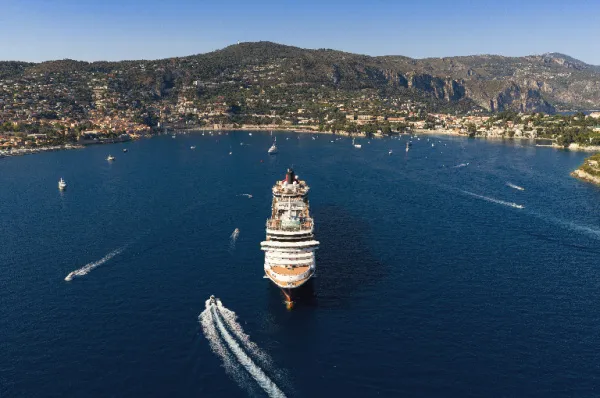 Luftaufnahme eines Cunard-Kreuzfahrtschiffes, das in einer sonnigen Bucht vor einer malerischen Küstenstadt im Mittelmeer ankert.