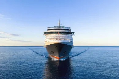 Eleganz auf See: Eine Cunard Queen auf majestätischer Fahrt Frontalansicht eines majestätischen Cunard Kreuzfahrtschiffes, das bei Sonnenschein auf dem offenen, blauen Meer fährt.