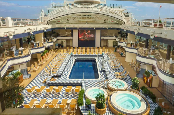 Eine Aufnahme des sonnigen Pooldecks auf einem Cunard-Schiff. In der Mitte befindet sich ein rechteckiger Swimmingpool, umgeben von zahlreichen Sonnenliegen auf einem blau-weiß karierten Deck.