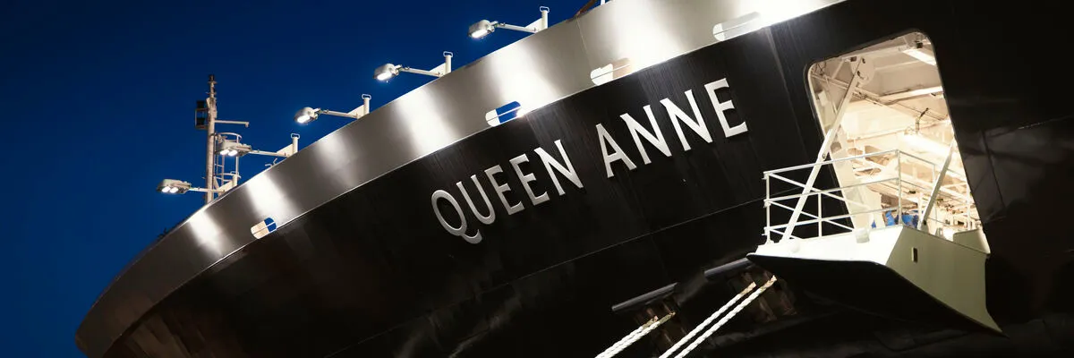 Eine Nahaufnahme bei Nacht, die den Rumpf des Cunard-Kreuzfahrtschiffs Queen Anne zeigt. Der Schiffsname „QUEEN ANNE“ ist in großen, von oben angestrahlten Buchstaben auf dem dunklen Rumpf zu sehen.