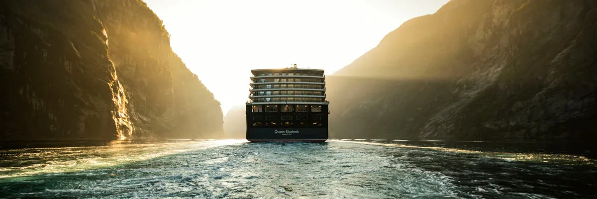 Eine Heckansicht des Cunard-Kreuzfahrtschiffs Queen Elizabeth, das bei Sonnenauf- oder -untergang durch einen engen Fjord fährt. Das Schiff entfernt sich, während die Sonne die steilen Felswände in ein goldenes Licht taucht.