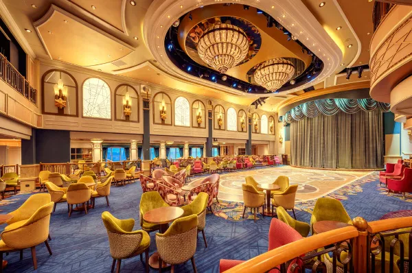 Eine Innenaufnahme des eleganten Queens Room Ballsaals auf einem Cunard-Kreuzfahrtschiff. Der Raum verfügt über eine große, hölzerne Tanzfläche in der Mitte, umgeben von Tischen und bequemen Sesseln in Senfgelb und Rosa.