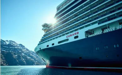 Eine Nahaufnahme der Seite und des Bugs eines Cunard-Kreuzfahrtschiffs, das durch einen Fjord oder eine bergige Küstenlandschaft fährt. Die Sonne scheint hell über der Brücke des Schiffes und spiegelt sich im ruhigen Wasser.