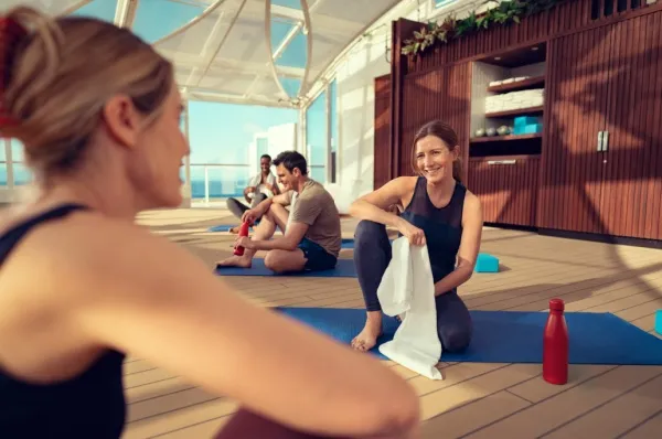 Frauen sitzen auf Yogamatten und unterhalten sich. In der Hand ein weißes Handtuch und auf dem Boden steht eine Trinkflasche.
