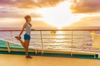 Eine Frau in Sportklamotten dehnt ihr Bein auf dem Oberdeck bei Sonnenuntergang, im Hintergrund das Meer.