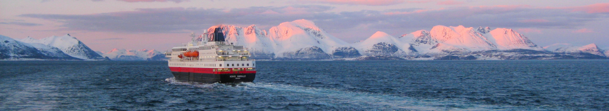 Das Hurtigruten-Schiff MS Kong Harald fährt auf blauem Meer vor einer verschneiten Gebirgskette in der Abendsonne.