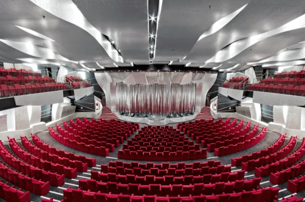 Eine weite Panoramaansicht des Pantheon Theatres auf der MSC Divina, gefüllt mit roten Samtsitzen auf mehreren Ebenen, die auf die große Bühne mit silbernem Vorhang blicken.