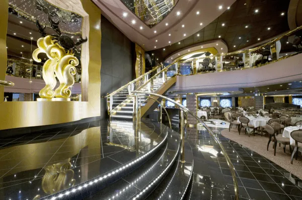 Der opulente Eingangsbereich eines Hauptrestaurants auf der MSC Divina mit einer breiten, beleuchteten Treppe, glänzenden schwarzen Böden und einer großen, goldenen Skulptur.