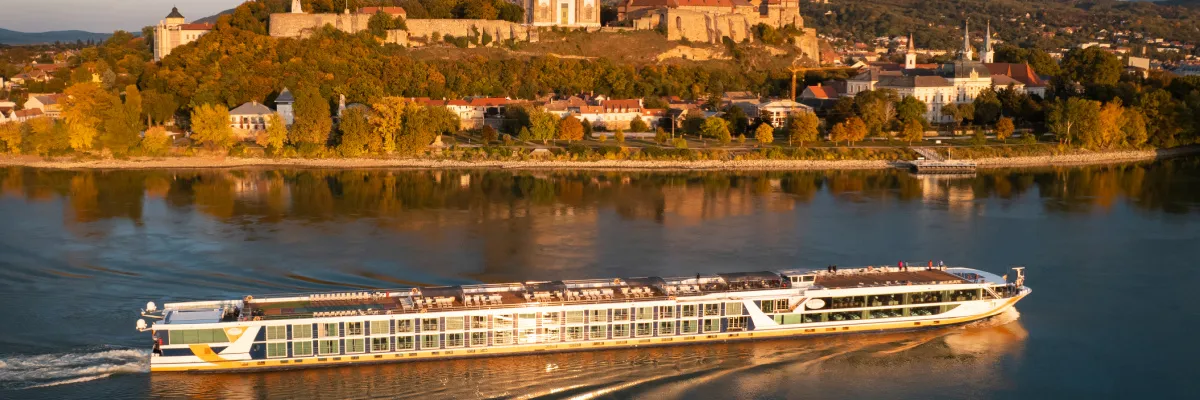 Ein 1A Vista Flusskreuzfahrtschiff fährt auf der Donau im Abendlicht vor der Kulisse der Burg und der Basilika von Esztergom in Ungarn.