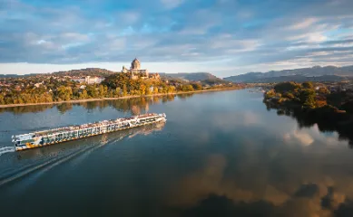 Ein langes Flusskreuzfahrtschiff fährt bei sonnigem Wetter auf einem breiten Fluss. Im Hintergrund ist eine Uferlandschaft mit einer Stadt und einer markanten Burg oder Kirche auf einem Hügel zu sehen, die sich im Wasser spiegelt.