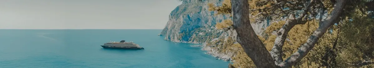 Eines der luxuriösen Schiffe von Explora Journeys vor einer felsigen Küste im Mittelmeer. Auf der rechten Seite steht ein herbstlicher Baum.