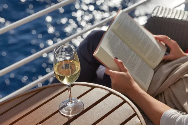 Eine Frau liest ein Buch und genießt ein Glas Weißwein auf dem Balkon der Deluxe Veranda Kabine auf einem der Regent Seven Seas Cruises Schiffe.