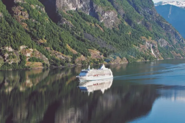 Die Seven Seas Yoyager im Geiranger Fjord in Norwegen. Die Felsen an der Klippe sind begrünt und das Wasser spiegelt das luxuriöse Kreuzfahrtschiff.