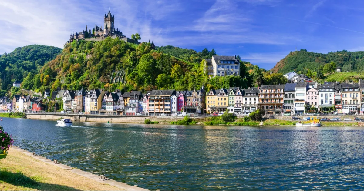 Eine Ortschaft mit bunten Häusern am Ufer eines Flusses, im Hintergrund thront eine Burg auf einem Berg unter blauem Himmel.