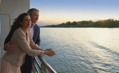 Ein Paar steht auf dem Balkon ihrer Kabine und blickt auf einen Fluss bei Sonnenuntergang.