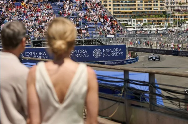 Ein Paar steht nah an der Formel 1 Rennstrecke beim Grand Prix de Monaco und schaut sich das Rennen an. Auf einem Banner ist das Logo von Explora Journeys.