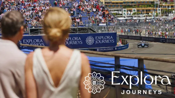 Ein elegantes Pärchen steht direkt an der Rennstrecke des Formal 1 Grand Prix in Monaco und schaut sich die Rennautos an. Auf einem Banner ist das Logo von Explora Journeys abgebildet.