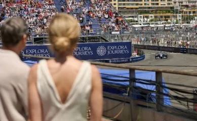 Kreuzfahrt zum Grand Prix de Monaco Ein elegantes Pärchen steht direkt an der Rennstrecke des Formal 1 Grand Prix in Monaco und schaut sich die Rennautos an. Auf einem Banner ist das Logo von Explora Journeys abgebildet.
