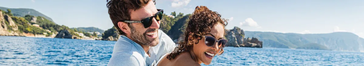 Bannerbild: Nahaufnahme eines herzlich lachenden Paares mit Sonnenbrillen auf dem Deck eines Kreuzfahrtschiffes, im Hintergrund tiefblaues Meer und eine sonnige Küstenlandschaft mit grünen Hügeln.